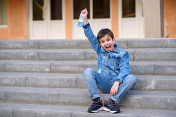 Happy boy sits on school stairs and takes off his protective mask