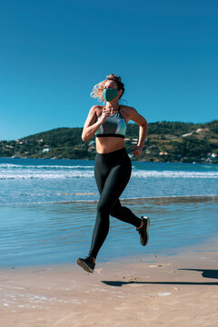 Beautiful athletic young woman running on the beach with green hills on the background wearing mask and sportswear on a sunny day and blue sky during covid-19 pandemic