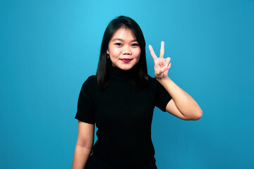 Portrait of Young beautiful asian women wearing black turtle neck shirt with blue isolated background,  Showing Peace Sign gesture concept