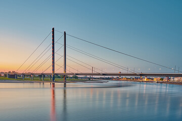 A cable-stayed bridge that leads across the Rhine. It is evening and the lighting of the bridge and the city of Düsseldorf are noticeable.    