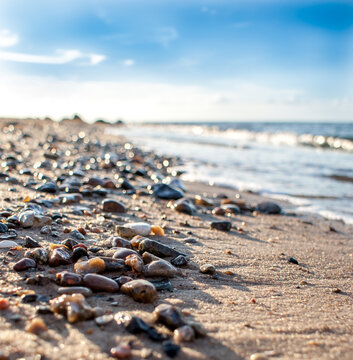 Vertical photo shows small stones at a sandy beach. The sky is blue, the sun is shining and in the right half of the picture is the sea. The picture was taken  from a low angel. 