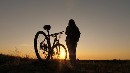 Hiker healthy young woman stands on a hill next to a bicycle, enjoying nature and sun. Free girl travels with a bicycle at sunset. concept of adventure and travel. lonely woman cyclist resting in park