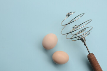 two chicken eggs and  manual hand egg beater on a blue background