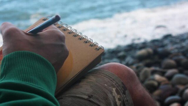 Man doing color pencil drawing in brown paper sketchbook by the sea