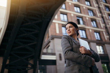 Bottom view portrait of an American business lady in a jacket holding a laptop in her hands and standing under a bridge