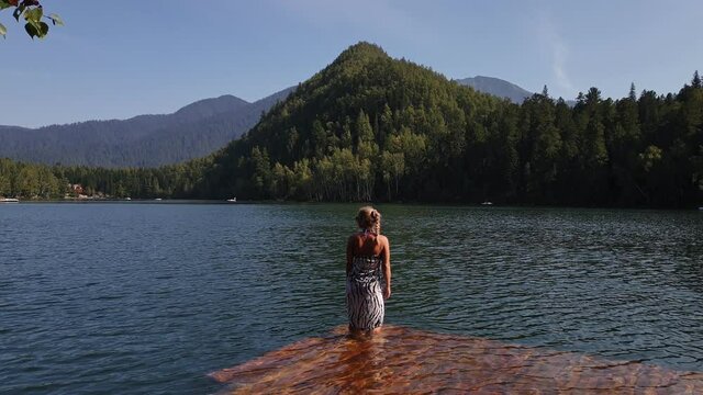 Woman walk on water on pier in sunglasses and a boho silk shawl. Girl rest on a flood wood underwater dock. The pavement is covered with water in the lake. In the background are mountain and a forest.