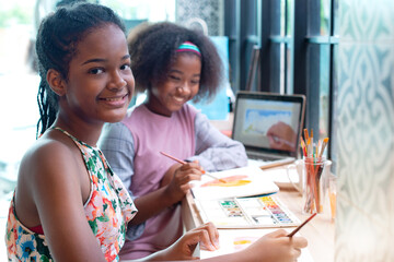 Two smiling African teen girls are drawing with watercolor and one is watching the camera