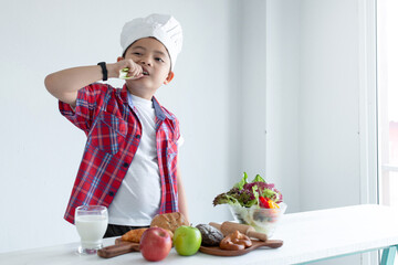 Asian kid wears a chef's hat and bites vegetables on a white background, enjoying and loving eating his vegetables