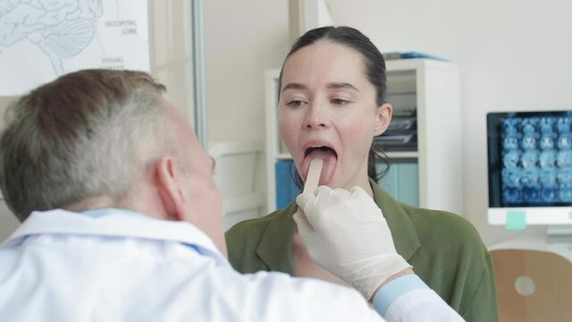 Back view of grey-haired male physician examining throat of young Caucasian woman with long dark hair sitting in front of him