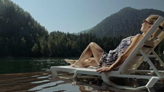 Woman lie on a sunbed in sunglasses and a boho silk shawl. Girl rest on a flood wood underwater pier. The pavement is covered with water in the lake. In the background are mountain and a forest.