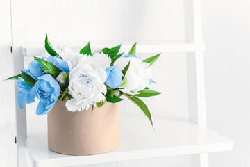 Box with beautiful peony flowers on shelf near white wall