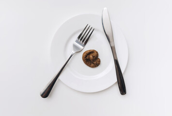 Plastic excrement of lies on a plate with fork and knife close-up on white tablecloths. The concept of an inept cook, fake news, a crappy restaurant, bad food, a taste of shit.Bad smelling joke.