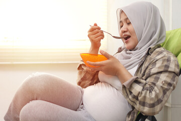 Happy Asian muslim pregnant mom sitting on deck chair and eating her healthy food for her baby