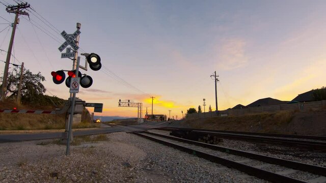 A railroad crossing at sunset.  The lights on the sign are flashing and a commuter train passes the camera.