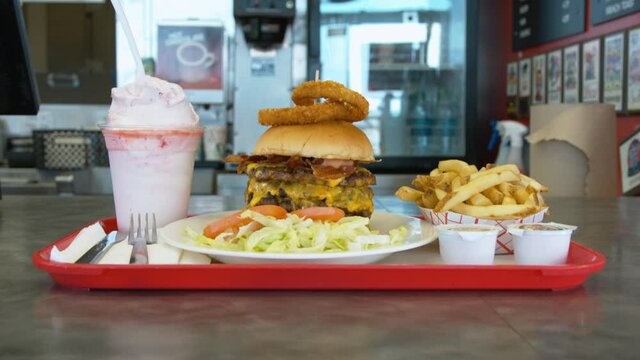 Woman employee at a burger joint pushes a full tray with the largest burger and fries and shake across the counter for the customer to pick up.