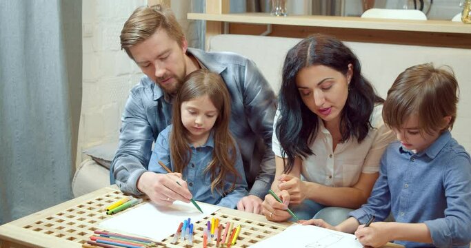 Happy creative family with two cute children drawing with colored pencils and markers on paper together sitting on the sofa at home.