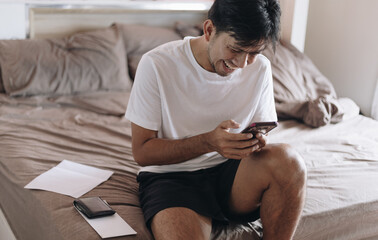 Young Asian man smiled and enjoyed while using the smartphone on the bed in the bedroom.