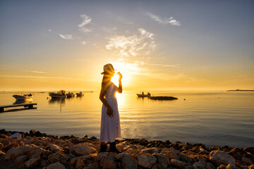 woman wearing white long dress with hat holding coffee cup standing on the beach island over sunrise morning sky with boats floating in the water background, Bahrain.