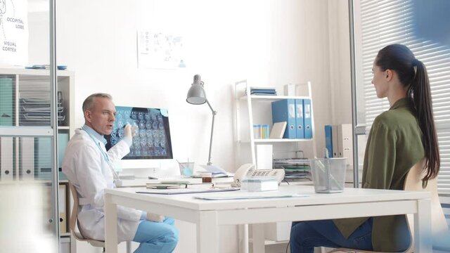 Medium shot of aged male doctor wearing medical overall showing x-ray image of brain to Caucasian brunette sitting in front of him and listening
