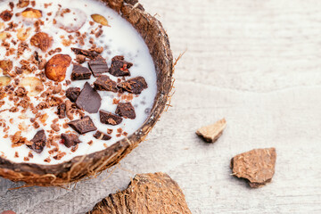  Coconut milk and granola made from oatmeal, nuts,dried fruits and grated chocolate in a coconut shell on a wooden background.Сoncept of healthy food and healthy desserts.Copy space for text