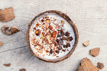  Coconut milk and granola made from oatmeal, nuts,dried fruits and grated chocolate in a coconut shell on a wooden background.Сoncept of healthy food and healthy desserts.Copy space for text