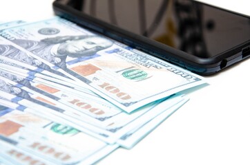Dollars and smartphone. Banknotes in denominations of 100 dollars lie on a white background on them, next lies a smartphone with a black screen. Top view and angle. Macro smartphone and dollars