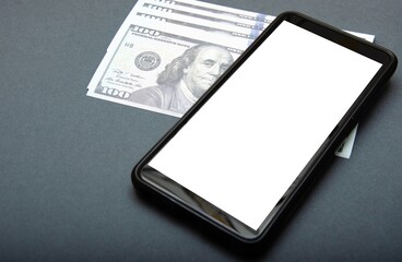 Dollars and smartphone. Denominations of 100 dollars are on a black background on them, next to them is a smartphone with a white screen. Top view and angle. Macro smartphone and dollars