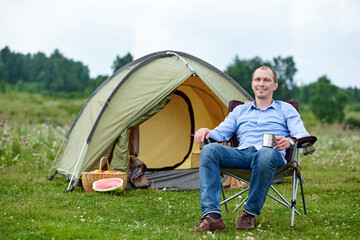 Naklejka na ściany i meble young man freelancer sitting on chair and relaxing in front of tent at camping site in forest or meadow. Outdoor activity in summer. Adventure traveling in national park. leisure, vacation, relaxation