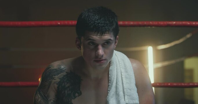 Close up of young Caucasian sweaty male guy boxer with tattoo and towel on shoulder sitting in corner of ring and looking down. Tired and upset sportsman having break. Loosing concept. Lost at fight.