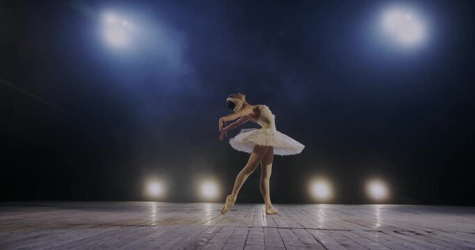 Beautiful Caucasian ballerina in white tutu and on pointe perfoming and mastering dance pas on stage in blue lights of lamps. Woman professional dancer of classical ballet training. Art concept.