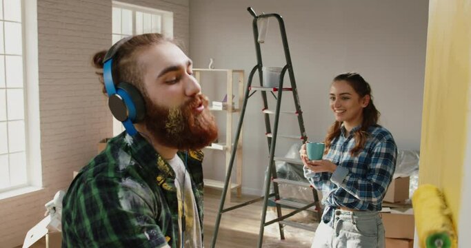 Cute newlywed couple renovating their home. Caucasian girl is offering a drink for her boyfriend who is painting a wall while listening to music - new life, love concept 4k footage