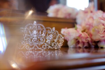A Golden crown of glittering stones lies on a wooden table in the room during the day. The bride's...
