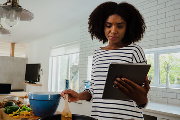 Beautiful mixed race woman looking at recipe on tablet while cooking in her kitchen at home