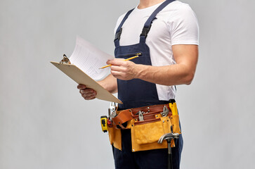 repair, construction and building - male worker or builder with clipboard, pencil and working tools on belt over grey background