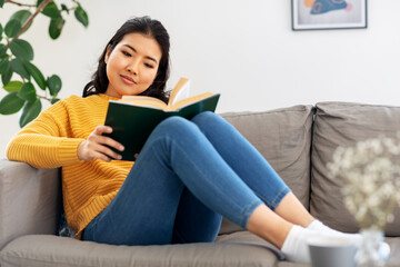 people and leisure concept - happy asian young woman in yellow sweater sitting on sofa and reading book at home