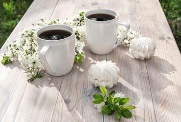 Two beautiful porcelain coffee cups with marshmallow dessert decorated with white cherry flowers on wooden table in spring garden in setting sun light.