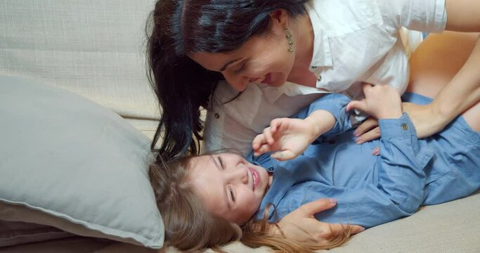 Cheerful family mother and daughter embracing and laughing lying on bed together. Happy young mom tickling her cute little daughter enjoying communication at home.