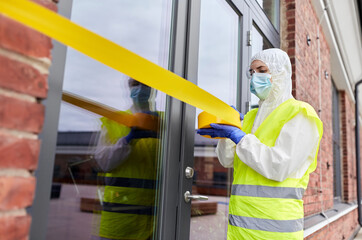 quarantine and pandemic concept - healthcare worker in protective gear or hazmat suit, medical mask, gloves and goggles enclosing building with caution tape outdoors