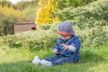 Naklejka na ściany i meble Cute little baby boy sitting outdoors is using smart phone. All potential trademarks are removed.