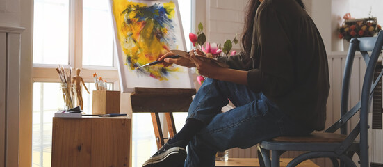Cropped shot view of Woman painter sitting on the floor in front of the canvas and drawing. Artist studio interior. Drawing supplies, oil paints, artist brushes, canvas, frame.
