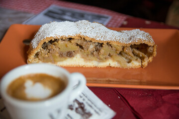 A slice of traditional Apple Strudel, powdered with sugar, served on a plate next to a cup of coffee