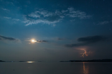 Lightning in the night starry sky with moon reflection over water. Incoming tropical thunderstorm over sea at the horizon. Inspirational background.