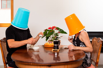 Young loving couple drinks coffee in the dining room with a bucket on their heads.