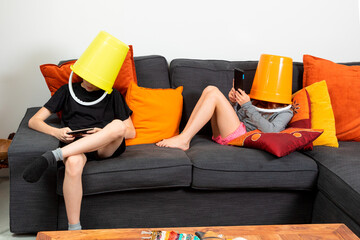 Brother and sister sitting on a sofa at home with a bucket on their heads playing with tablets