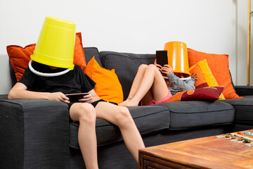 Brother and sister sitting on a sofa at home with a bucket on their heads playing with tablets