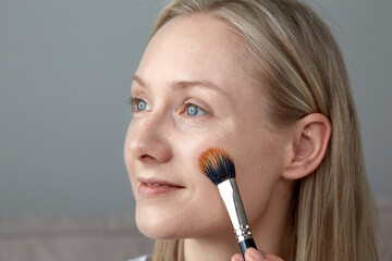 A beautiful young blonde with bright blue eyes paints her face with a brush.