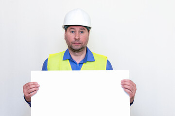 Close-up Engineer man wearing a helmet and reflector vest, standing and shows blank  billboard. White studio wall.