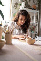 Beautiful young brunette girl in blue apron making ceramic and pottery tableware at the workshop. She using special handtool to cut out shapes from soft stuff while working over new item.
