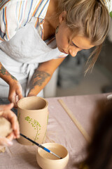 Fototapeta na wymiar Smiling beautiful girl in blue apron holding rolling pin and happily working with clay at pottery class studio. male potter master rolling up the clay on table with ceramic products.