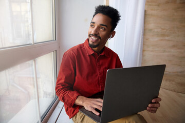 Positive young pretty short haired bearded brunette man with dark skin keeping laptop on his knees while sitting on windowsill and looking gladly out of window
